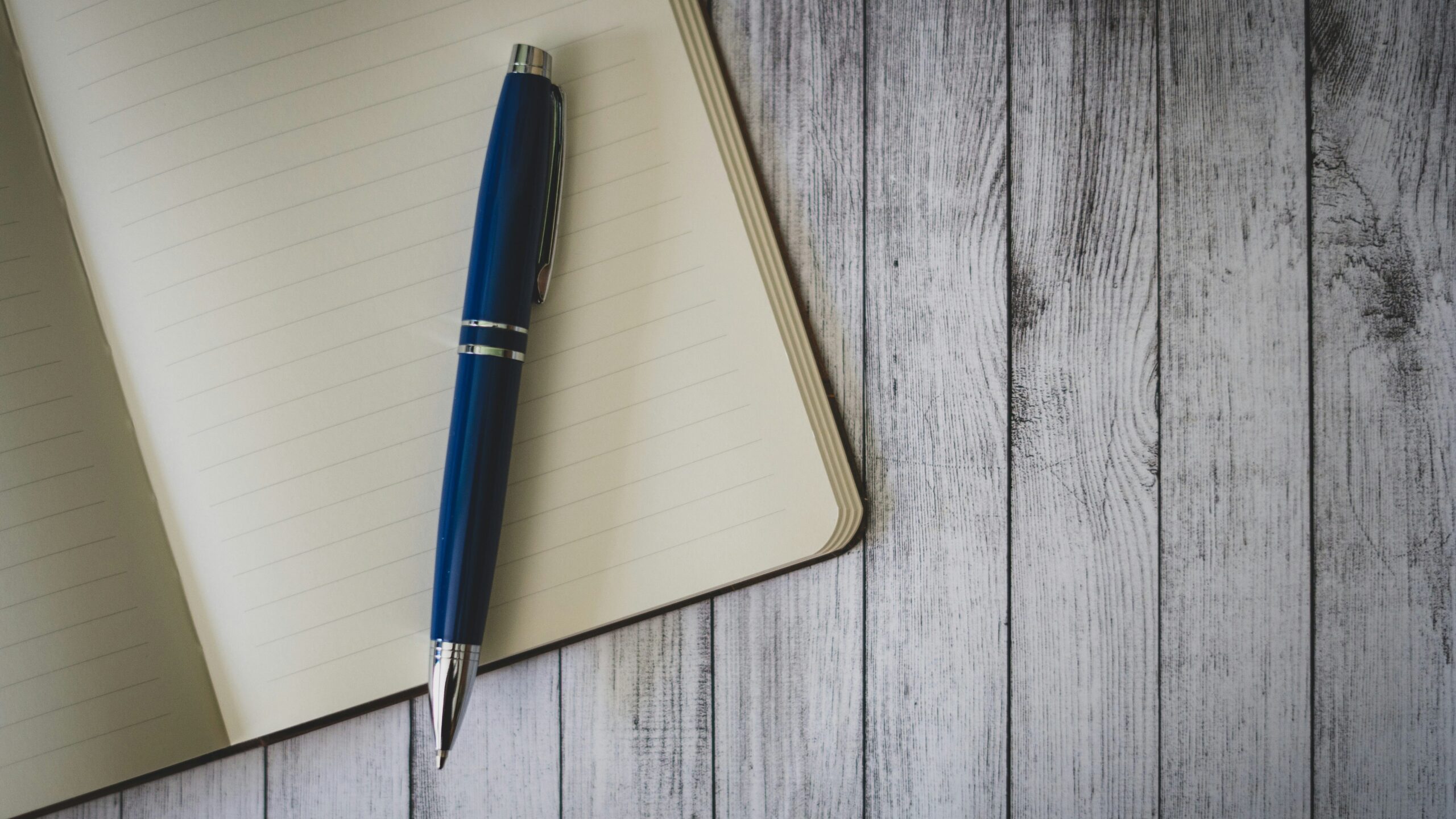 A top-down photo of a blue ballpoint pen on an open notebook on a grey wood table